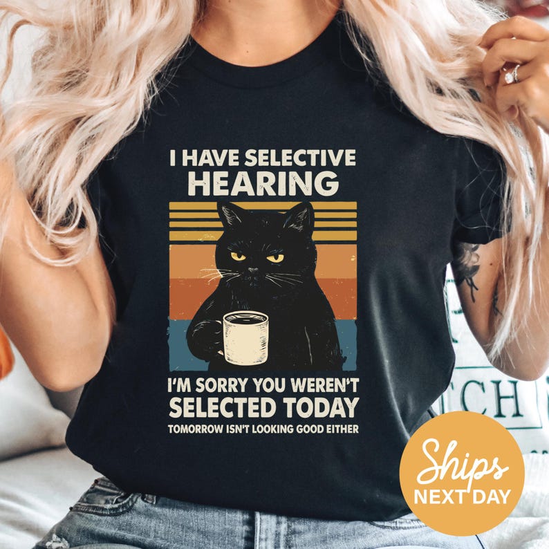 A person wearing a black t-shirt with a graphic design depicting a black cat holding a coffee cup and a humorous caption.