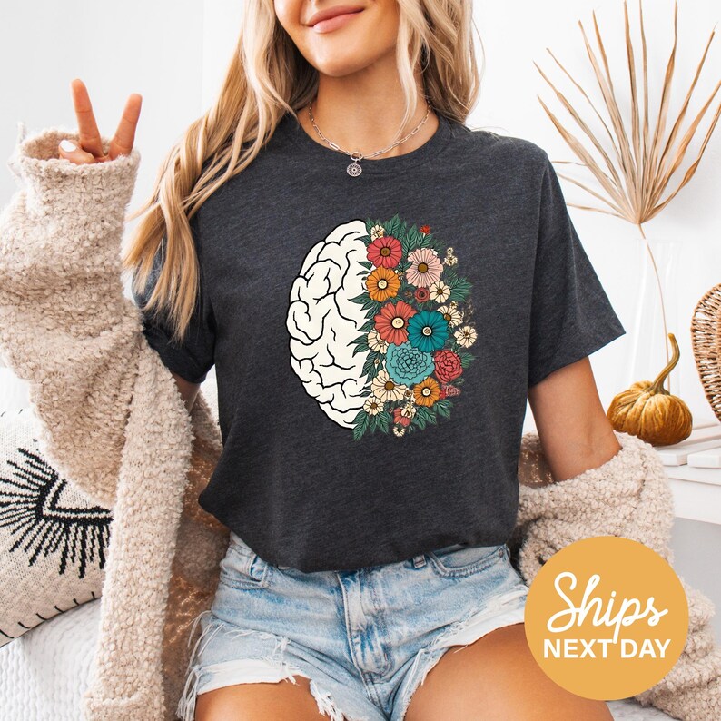 A woman wearing a gray t-shirt with a graphic of a brain surrounded by colorful flowers, making a peace sign gesture. She is also wearing denim shorts and a beige cardigan.