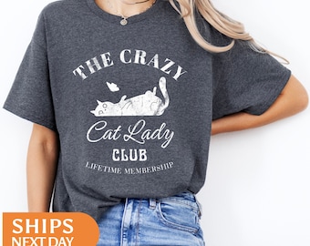 May include: Dark gray t-shirt with the text "THE CRAZY Cat Lady CLUB" and a graphic of a cat. The shirt also has the words "LIFETIME MEMBERSHIP" printed below the cat graphic. The shirt is paired with blue jeans.