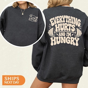 May include: A dark charcoal gray crewneck sweatshirt featuring a small graphic on the front left chest of a tired emoji holding a fork. The back displays a large graphic with a barbell and the text Everything Hurts And I'm Hungry in cream.