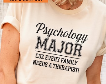 May include: A cream colored t-shirt with black text that reads "Psychology Major Cuz Every Family Needs A Therapist!"