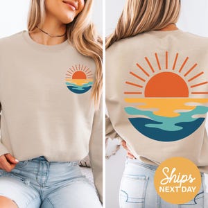 May include: Beige crewneck sweatshirt featuring a circular graphic of a sunset over water. The design includes an orange sun, yellow and blue water, and is repeated on the front and back. The sweatshirt is a neutral color, suitable for casual wear.