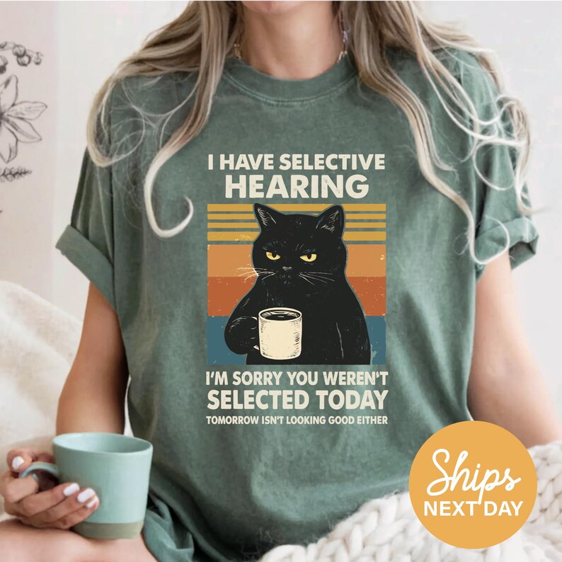 A woman wearing a green t-shirt with a graphic design of a black cat holding a coffee cup and text that reads "I HAVE SELECTIVE HEARING I'M SORRY YOU WEREN'T SELECTED TODAY TOMORROW ISN'T LOOKING GOOD EITHER".