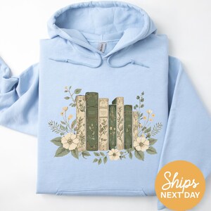 A light blue hoodie with a floral design featuring a row of books surrounded by flowers and leaves.