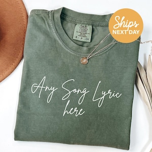 A green t-shirt with the text "Any Song Lyric here" printed on it, along with a necklace and a brown hat.