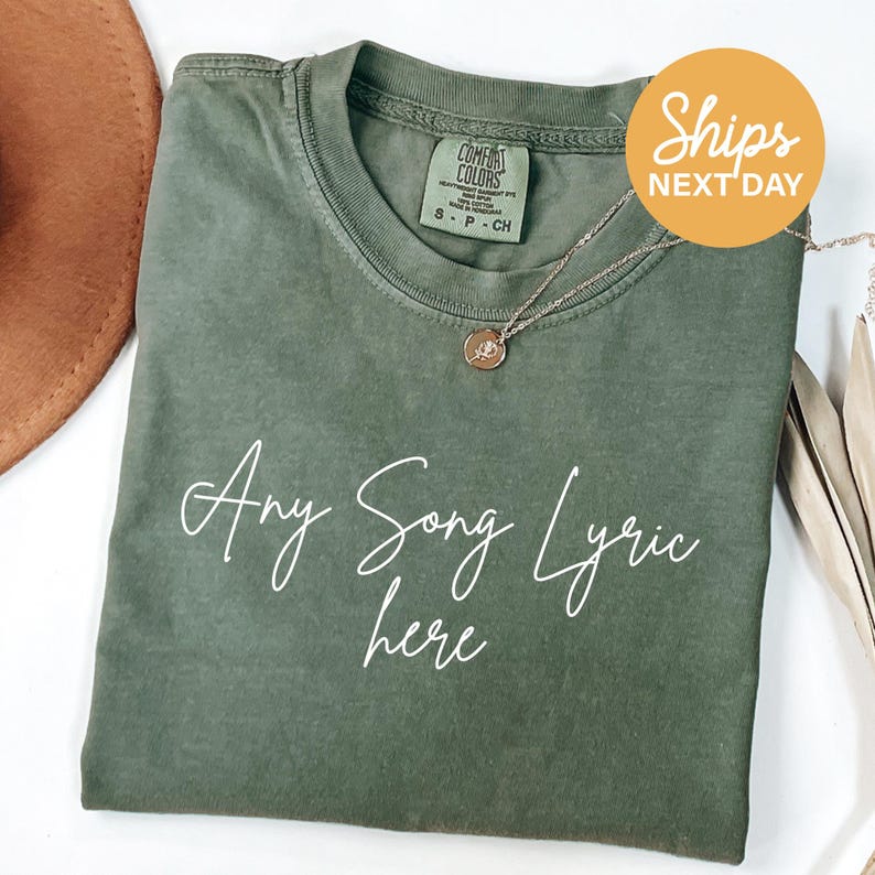 A green t-shirt with the text "Any Song Lyric here" printed on it, along with a necklace and a brown hat.