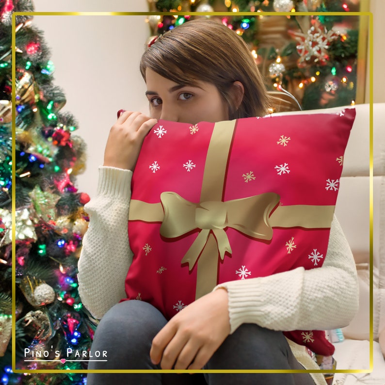 May include: A red and gold throw pillow with a gift box design and a white snowflake pattern. The pillow is being held by a person in front of a Christmas tree.