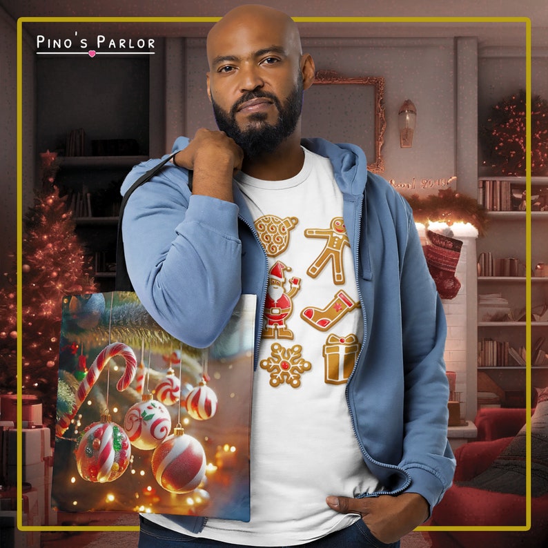 May include: A white t-shirt with a festive design featuring gingerbread cookies, including a snowman, a stocking, a gingerbread man, and a present. The shirt is being held by a person wearing a blue hoodie. The t-shirt is also being used as a tote bag with a Christmas tree and ornaments printed on it.