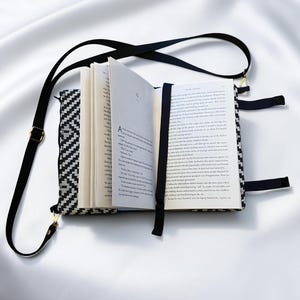 Könnte beinhalten: Ein offenes Buch mit einem schwarz-weiß gemusterten Einband und einem schwarzen Riemen. Das Buch wird von einem schwarzen Lesezeichen aus Band gehalten. Ein verstellbarer schwarzer Schulterriemen ist mit goldfarbener Hardware an der Hülle befestigt.