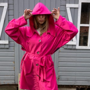 May include: A bright pink raincoat with a hood and a belt. The raincoat has a button closure and two pockets.