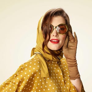 May include: A woman wearing a yellow polka dot shirt with a matching yellow headscarf and brown gloves. She is wearing large, round, gold-rimmed sunglasses.