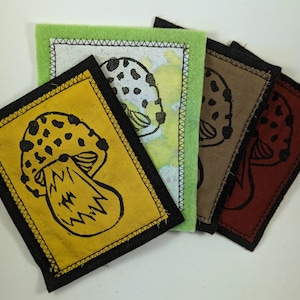 Peut inclure: Quatre patchs en tissu avec un motif de champignon noir. Les patchs sont de différentes couleurs : jaune, vert, marron et rouge. Chaque patch a une bordure noire cousue.