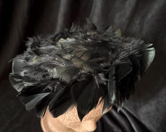 May include: A black feathered hat, likely a fascinator or headpiece, is the focal point. The hat is densely covered in dark feathers, creating a textured, voluminous appearance. The feathers have a slight sheen, and the hat is set against a dark background.