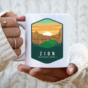 May include: White ceramic mug with a green, yellow, and orange graphic of a mountain range and the text "ZION NATIONAL PARK".