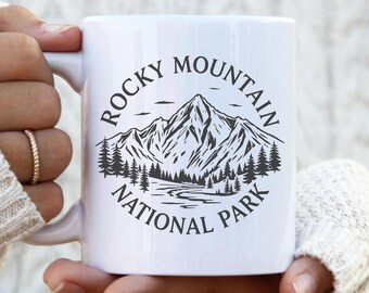 May include: White ceramic mug featuring a black and white graphic of a mountain range, trees, and the text "Rocky Mountain National Park" in a circular design. The mug has a white handle and is held by a person.