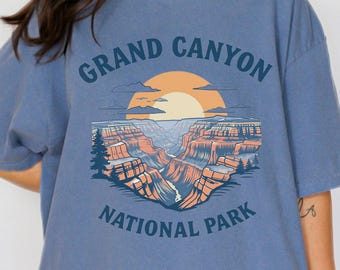 May include: Blue t-shirt with a graphic of the Grand Canyon National Park. The design features a sunset, canyon landscape, and the text "GRAND CANYON NATIONAL PARK" in navy blue.