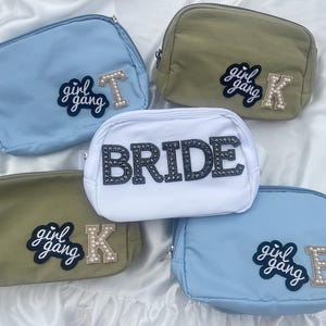 May include: Five different cosmetic bags with various colors and designs. The bags are made of fabric and have a zipper closure. The bags have different patches on them, including "girl gang" and "bride".
