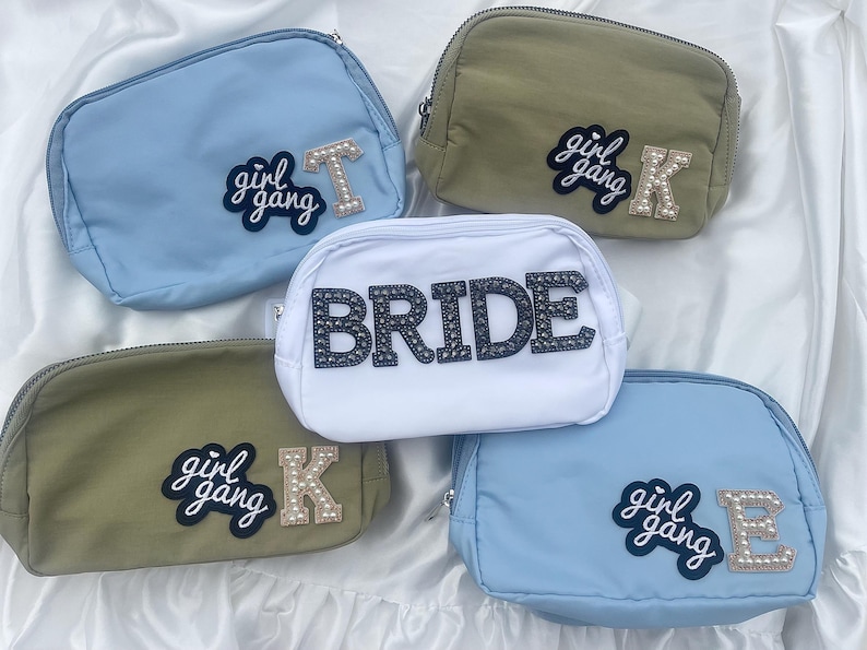 May include: Five different cosmetic bags with various colors and designs. The bags are made of fabric and have a zipper closure. The bags have different patches on them, including "girl gang" and "bride".