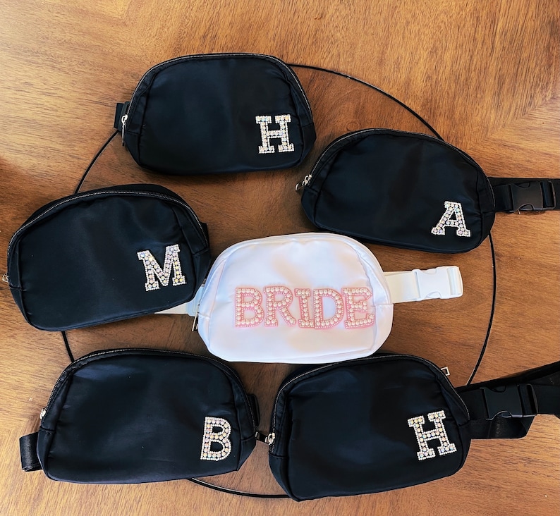 May include: Six black fanny packs with rhinestone letters. The letters spell out "BRIDE" and "M, A, H, H".