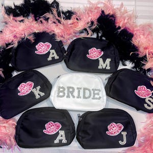 May include: Set of seven black cosmetic bags with zipper closures. Each bag has a different rhinestone letter and a pink lip design. One bag is white with the word "BRIDE" in rhinestones.