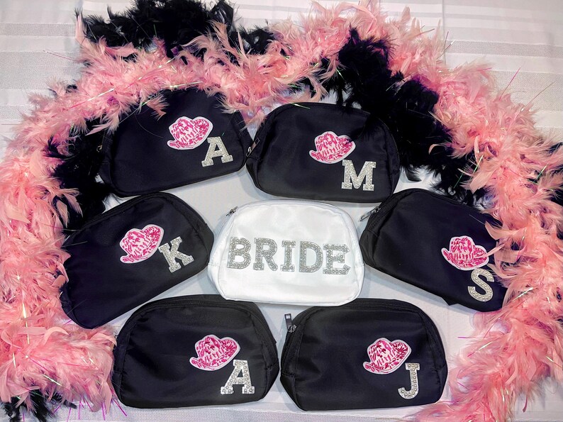 May include: Set of seven black cosmetic bags with zipper closures. Each bag has a different rhinestone letter and a pink lip design. One bag is white with the word "BRIDE" in rhinestones.