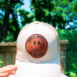 Pode incluir: Um bon&eacute; de caminhoneiro de malha branca e marrom com um remendo de couro marrom que diz "Don't Worry Be Happy" com um rosto sorridente.