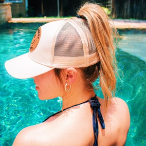 Pode incluir: Uma mulher com cabelo comprido e loiro est&aacute; usando um bon&eacute; de caminhoneiro de malha bege com um remendo de couro marrom que diz "smiley" em letras brancas. Ela est&aacute; em uma piscina com &aacute;gua azul.