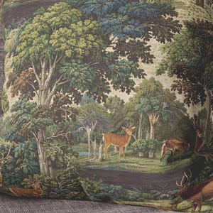 May include: A square throw pillow featuring a detailed tapestry-style print of a forest landscape. The scene depicts multiple deer standing and resting among lush green trees, foliage, and a small stream in earthy tones of brown and green.