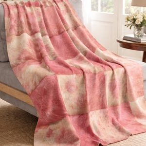 May include: A soft throw blanket featuring a patchwork design of alternating square panels in shades of dusty rose pink and cream. The fabric displays a subtle, mottled tie-dye effect across the surface, creating a textured appearance.