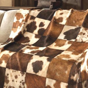 Peut inclure: Un plaid rectangulaire présentant un motif en patchwork composé de panneaux carrés. Le design alterne entre des sections marron foncé unies et des sections crème ornées de taches et de textures mouchetées rappelant la peau de vache.