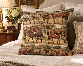 May include: A square throw pillow featuring a woven tapestry design of horses and riders in a rural setting. The scene depicts multiple brown and grey horses, people in red jackets, and stone buildings with green foliage on a beige background.