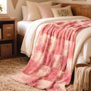 Op de afbeelding: Een pluche plaid met een ruitpatroon van afwisselende oudroze en crèmekleurige vierkanten met een zachte, getextureerde afwerking. De deken is gedrapeerd over een bed met wit beddengoed, gestreepte kussens en een kussen met bloemenprint.