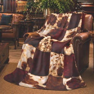 May include: A plush throw blanket featuring a patchwork design of faux cowhide patterns in shades of deep burgundy, dark brown, and cream. The blanket is draped over a brown leather armchair, showcasing a soft, textured surface with varied patches.