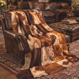 May include: A patchwork throw blanket with a cowhide design in shades of brown and tan. The blanket is draped over a brown leather armchair, showcasing its texture and pattern. The throw adds a rustic and cozy element to the room's decor.