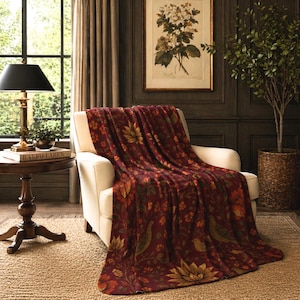 Può includere: Una coperta bordeaux con un motivo floreale e di uccelli drappeggiata su una poltrona bianca. La coperta presenta accenti dorati, arancioni e verdi. La scena è ambientata in una stanza con una parete con pannelli in legno scuro, un tavolino rotondo e una lampada da terra.