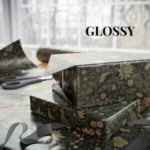 May include: Two gift boxes wrapped in glossy black paper with a floral and bird pattern. The word "GLOSSY" is printed in black. Scissors and a roll of wrapping paper are visible in the background, along with a gray ribbon.