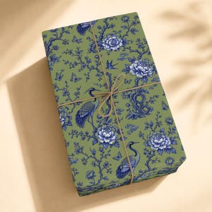 May include: A rectangular gift box wrapped in olive green paper featuring a repeating blue pattern of cranes, blooming peonies, butterflies, and branches. The package is tied with a thin, light brown twine string centered in a cross pattern.