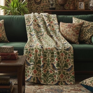 Pode incluir: Uma manta creme com um padrão repetido de pássaros azuis, flores vermelhas e folhagem verde, drapeada sobre um sofá verde. Almofadas decorativas com padrões semelhantes também são visíveis. O ambiente é vintage e acolhedor.