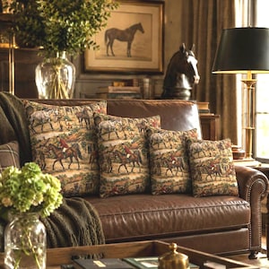 May include: Four square throw pillows featuring a woven tapestry pattern of horses and riders in a countryside setting. The pillows are arranged in a row on a brown leather sofa, displaying a repeating scene of figures on horseback.