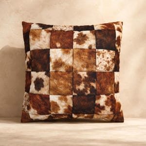 May include: A square throw pillow featuring a patchwork design composed of individual squares of cowhide. The pattern displays a variety of natural textures and colors, including shades of dark brown, light tan, cream, and white in mottled spots.