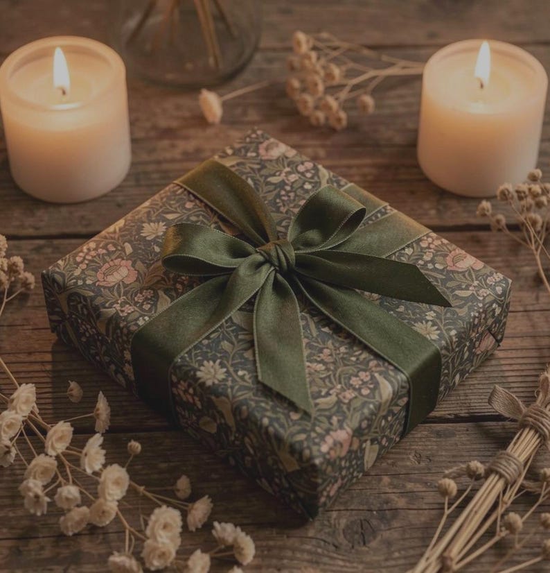 May include: A wrapped gift box with a floral pattern in shades of green, pink, and brown, tied with a dark green velvet ribbon. Two lit candles and dried flowers are arranged on a wooden surface, creating a warm, inviting scene.