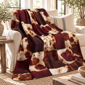 Puede incluir: Una manta de felpa con un diseño de patchwork de paneles de color rojo burdeos intenso que se alternan con patrones de manchas de color crema y marrón inspirados en la piel de vaca. La tela tiene una apariencia suave y texturizada con una disposición rectangular repetitiva.