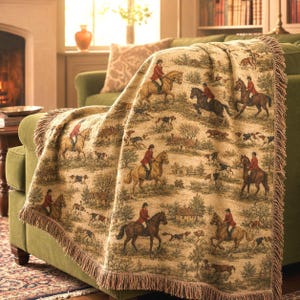 May include: A woven throw blanket featuring a repeating equestrian fox hunt scene with riders in red coats on horseback and hunting dogs across a tan background. The blanket has a textured fabric finish and decorative fringe along the edges.