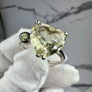 May include: A silver ring featuring a large, heart-shaped, pale yellow gemstone. A smaller, round, green gemstone is set beside the heart. The ring is held by a gloved hand, showcasing the jewelry's design and color.