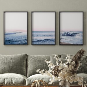 May include: Three framed ocean wave prints. Each print features a view of the ocean with blue waves and a pink and white sky. The frames are black and the prints are displayed above a gray sofa.