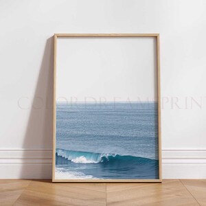 May include: A framed photograph of a blue ocean wave crashing against the shore. The wave is white and foamy, and the ocean is a deep blue. The photo is framed in a light wood frame.