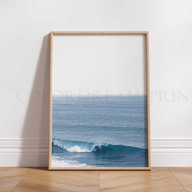 May include: A framed photograph of a blue ocean wave crashing against the shore. The wave is white and foamy, and the ocean is a deep blue. The photo is framed in a light wood frame.