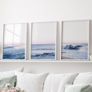 May include: Three framed ocean wave prints. Each print features a different view of the ocean with blue water and a pink and blue sky. The frames are white and the prints are displayed on a white shelf.