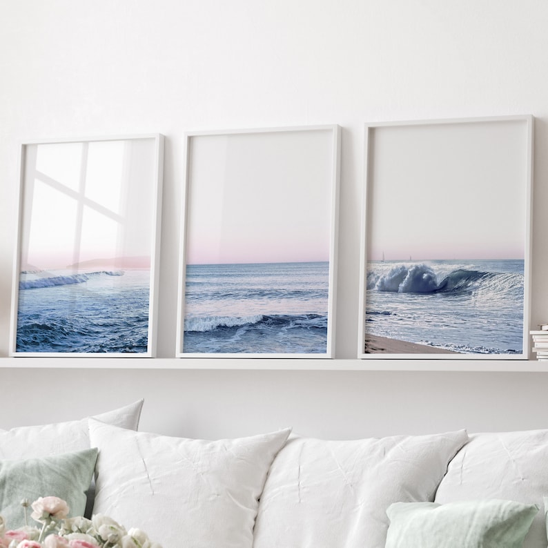 May include: Three framed ocean wave prints. Each print features a different view of the ocean with blue water and a pink and blue sky. The frames are white and the prints are displayed on a white shelf.