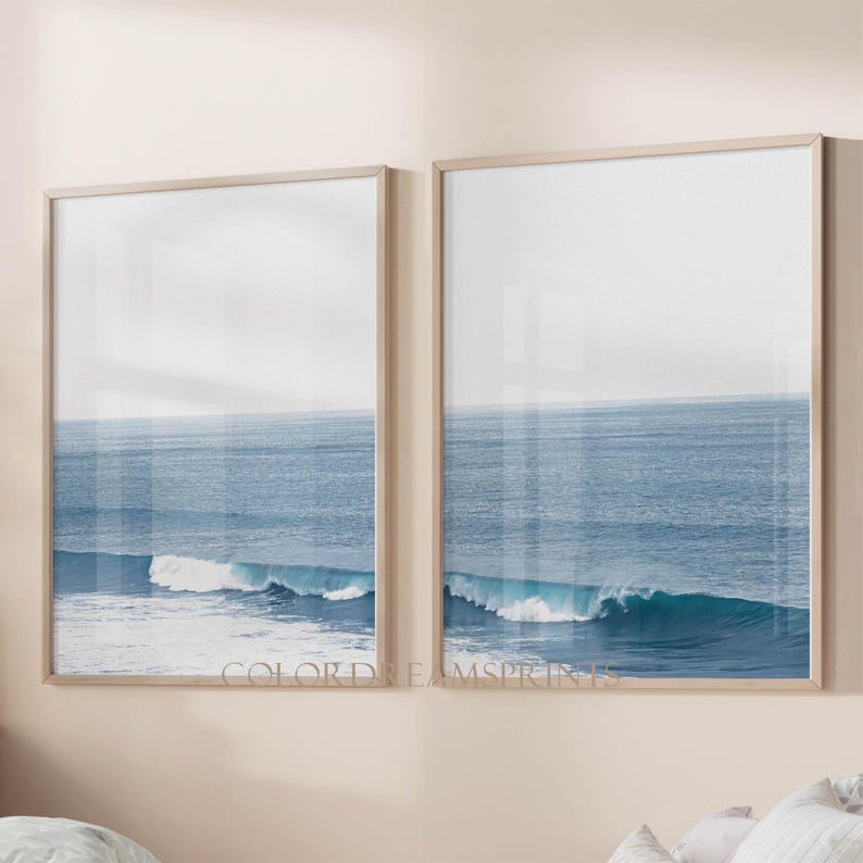 May include: Two framed prints of a blue ocean with white waves crashing on the shore. The prints are in a light wood frame and are hung on a light beige wall.